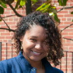 Sherine A woman with curly hair wearing a blue jacket stands outside in front of a brick wall and a tree, capturing the serene environment as she experiences Saint Joseph's College of Maine campus. Saint Joseph's College of Maine