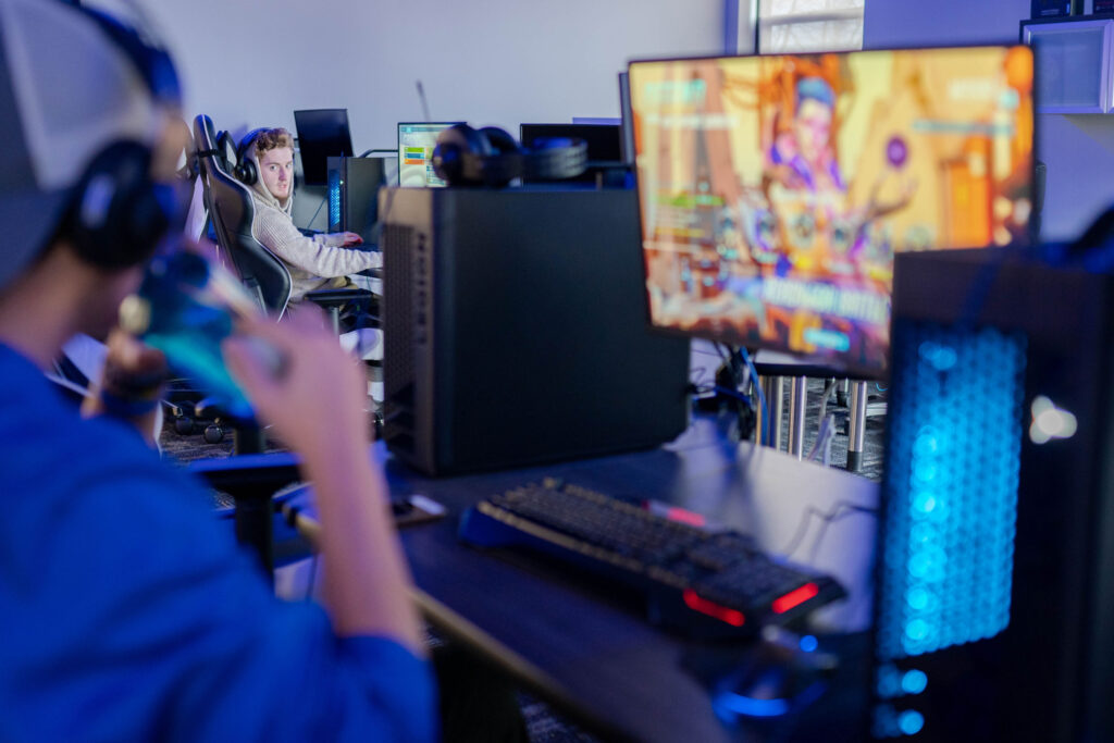 People are playing video games on desktop computers in a gaming setup, with one person in focus and another blurred in the foreground. The screen displays a colorful game, creating an immersive experience reminiscent of campus life and admissions events. Saint Joseph's College of Maine