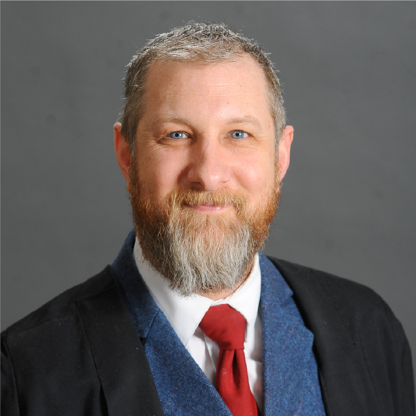 Smiling man with gray hair and beard, PhD, Jake Thibault, wearing a blue suit, white shirt, red tie, and black jacket, posed against a plain gray background. Saint Joseph's College of Maine