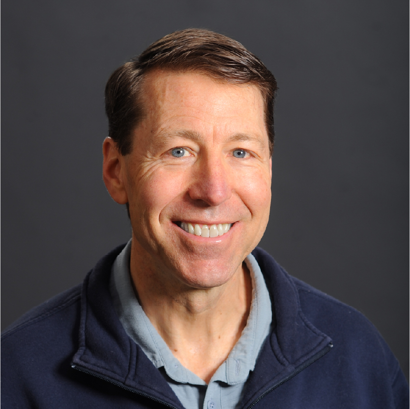 Steven L. Bridge, a man with short brown hair, wearing a blue fleece jacket over a light blue shirt, is smiling in front of a plain gray background. Saint Joseph's College of Maine