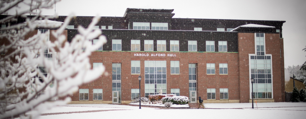 The image shows the exterior of Harold Alfond Hall, a multi-story brick building with large windows, covered in snow. A snowy tree branch is visible in the foreground, inviting you to experience Saint Joseph's College of Maine campus in its serene winter beauty. Saint Joseph's College of Maine
