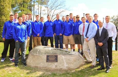 A group of people, most in matching blue jackets, are standing outdoors around a rock with a plaque on it. Some wear casual attire while others are dressed in business attire. The trees and clear sky provide a beautiful backdrop for the Plaque Dedication Ceremony at Clark’s Court. Saint Joseph's College of Maine