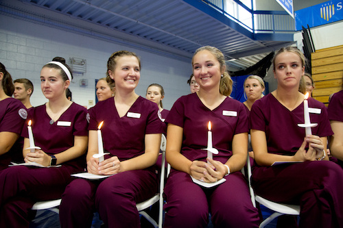 A group of nursing students from the Class of 2020, dressed in maroon scrubs, sit in a row holding lit candles during their Nursing Lighting Ceremony. Saint Joseph's College of Maine