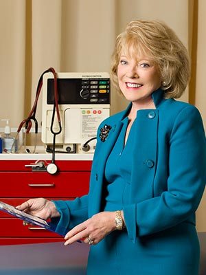 A smiling woman in a teal suit holds a clipboard in a medical office at Saint Joseph's College, with a defibrillator and medical equipment in the background. Saint Joseph's College of Maine