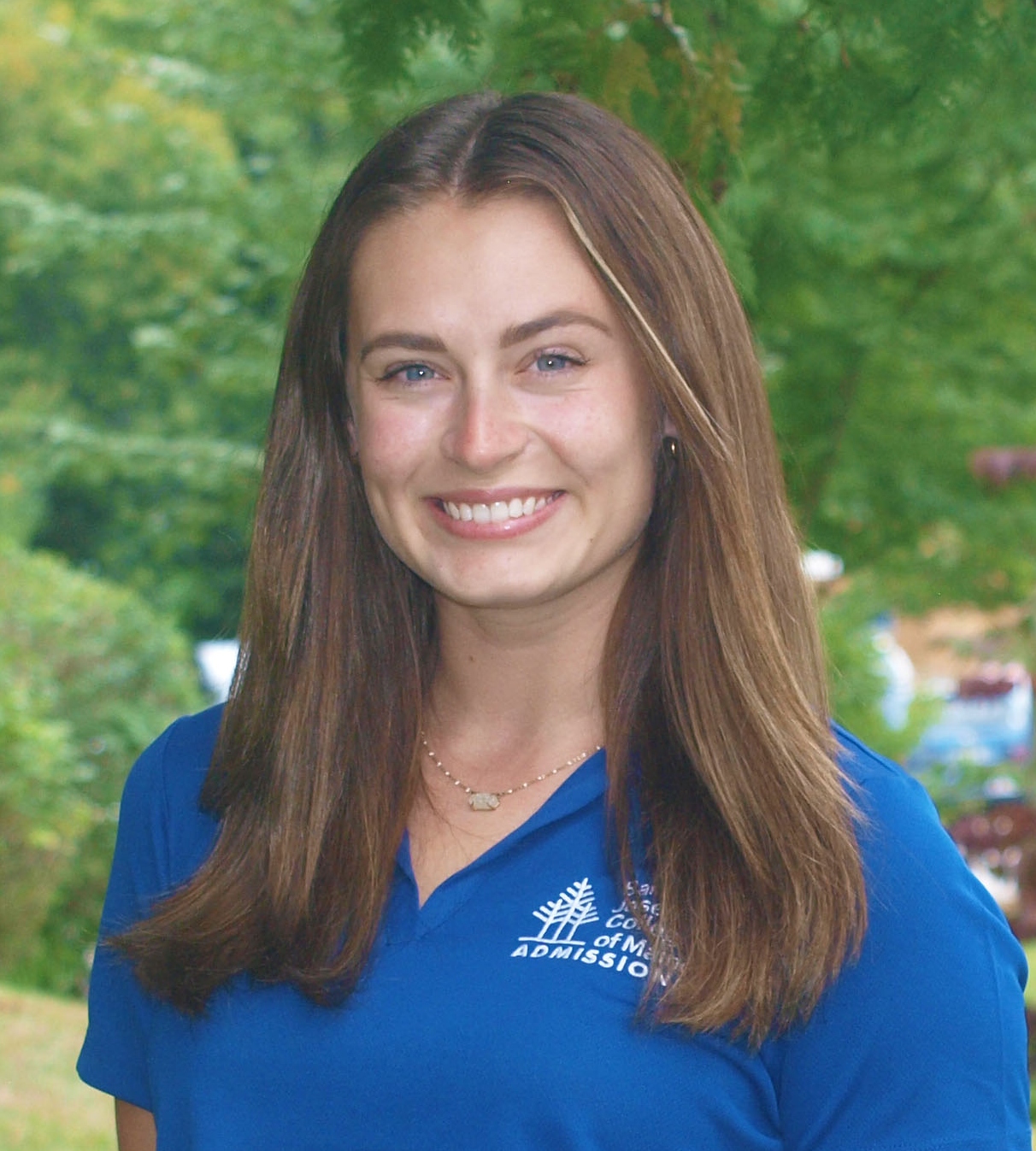 Abby-OBrien A person with long brown hair smiles at the camera. They are wearing a blue polo shirt with a college logo. The background is a blurred outdoor setting with greenery. Saint Joseph's College of Maine