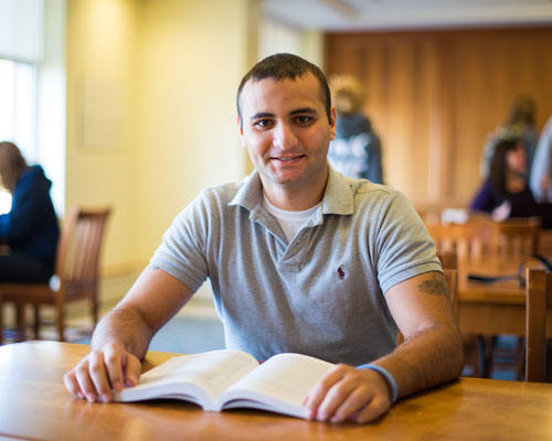 A man sits at a library table with an open book in front of him, smiling at the camera. People in the background, immersed in their own books, are also engaged in studying. His worn boots peek out from under the table, suggesting he’s been here for a while. Saint Joseph's College of Maine