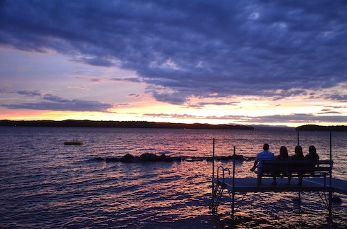 Four alumni sit on a bench at the end of a dock, overlooking a lake during a colorful sunset with purple and orange hues in the sky, reminiscing about their college reunion. Saint Joseph's College of Maine