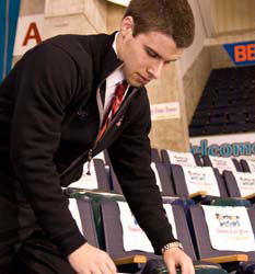A man in a black jacket, potentially guiding students through their sports management internships, is methodically arranging items on a row of stadium seats. Saint Joseph's College of Maine