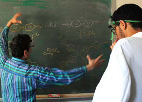 A professor gestures towards a chalkboard with chemical structures while a student in a lab coat and goggles observes thoughtfully, immersing themselves in the rich laboratory experience. Saint Joseph's College of Maine