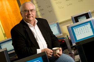 A man with short gray hair and glasses, dressed in a black blazer and white shirt, holding a coffee cup, is seated in a computer lab with multiple monitors, embodying the spirit of new journalism amidst the hum of conversations that feel like people talking at each other. Saint Joseph's College of Maine