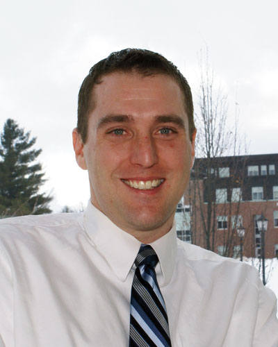Michael Bolanz, the Director of Alumni Relations, is smiling in front of a window with trees and a building in the background, wearing a white shirt and striped tie. His friendly demeanor embodies his commitment to team leadership. Saint Joseph's College of Maine