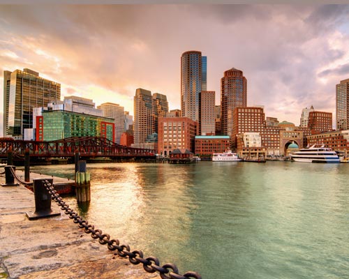Waterfront cityscape featuring high-rise buildings and boats docked at the harbor. A chain barrier lines the stone pathway along the water, where a regional alumni chapter gathers for their annual reunion. The sky shows hints of sunset or sunrise colors, adding to the picturesque scene. Saint Joseph's College of Maine