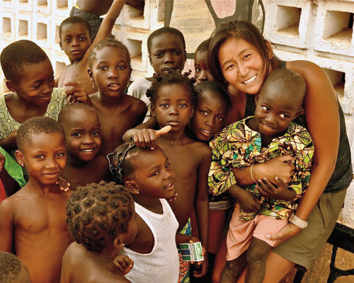 A smiling woman, living the mission, is surrounded by a group of children in an outdoor setting. She is holding one of the children, and all are looking at the camera, serving others with warmth and joy. Saint Joseph's College of Maine