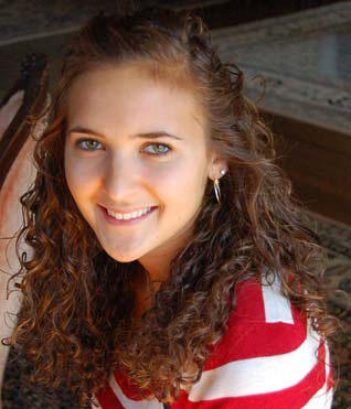 Young woman, Caitlyn Smith, with long curly hair and green eyes, smiling at the camera. She is wearing a red and white striped shirt and hoop earrings—class of '11. Saint Joseph's College of Maine