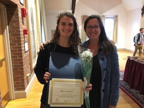 Two women indoors, smiling. The woman on the left is holding a certificate and a flower bouquet, having just received the Heart & Soul Student Award. The woman on the right stands beside her with her arm around her shoulder. Saint Joseph's College of Maine