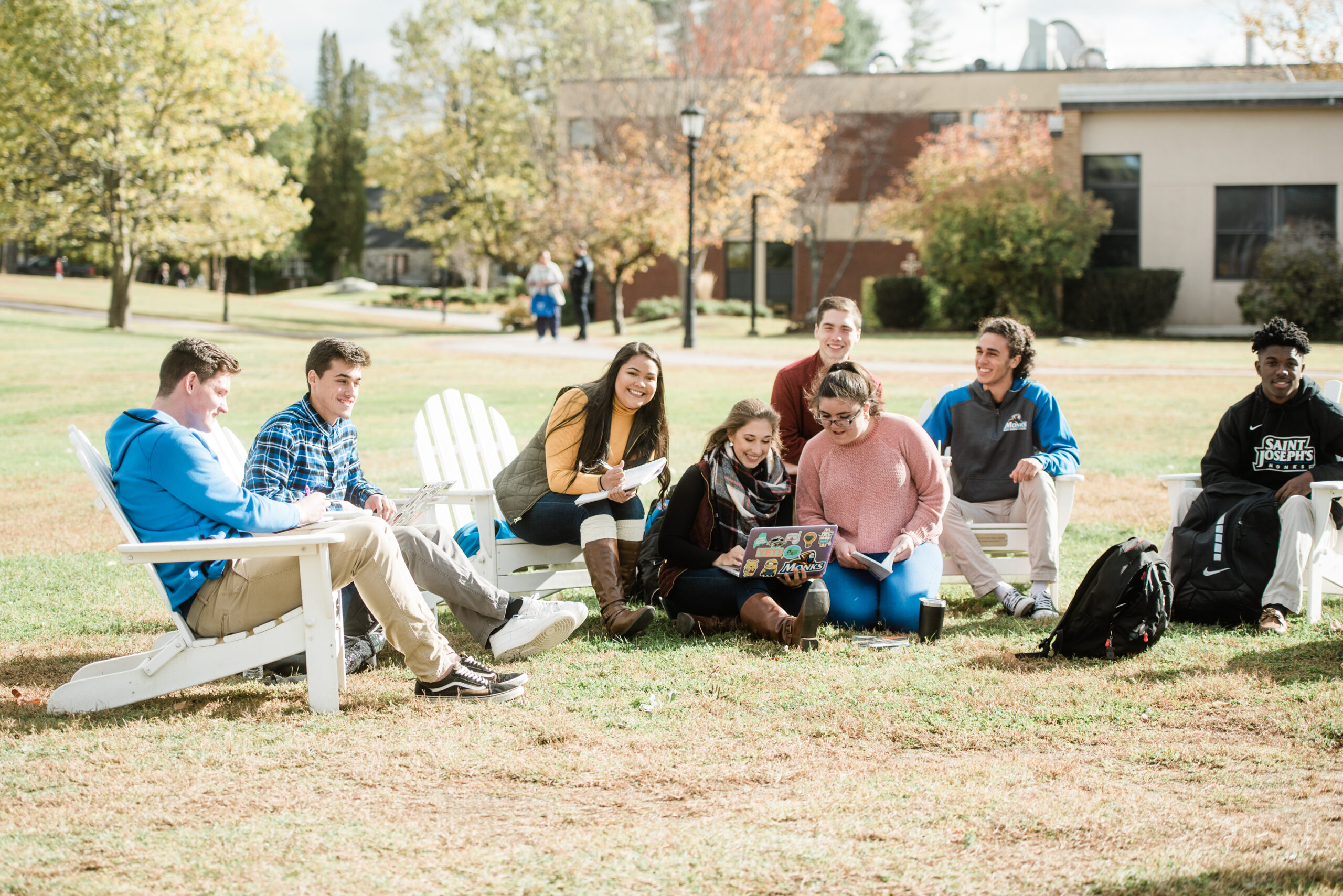 A group of people sit on Adirondack chairs and on the grass in a park-like setting with buildings and trees in the background. Some are looking at a laptop, likely discussing their achievements from the Fall 2019 semester, perhaps even celebrating those who made the Dean’s List. Saint Joseph's College of Maine