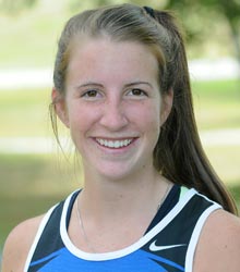 A woman with long brown hair in a ponytail smiles while wearing a blue and black athletic tank top, standing outdoors with blurred greenery in the background, as if she's just finished reading some uplifting campus news. Saint Joseph's College of Maine
