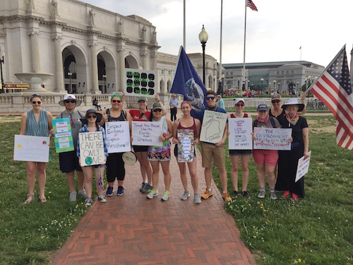 A group of people stands outside a building, holding various signs with messages on science and environmental issues. Among them are students from Saint Joseph's College, wearing casual clothes, some with hats and sunglasses. Their rally supports the Paris Climate Agreement. Saint Joseph's College of Maine