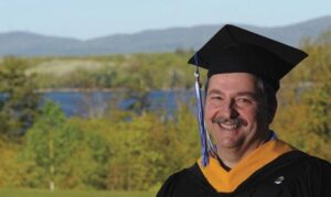 David Stamm, in his own words, stands proudly outdoors in graduation attire. He wears a black cap and gown with a gold-colored sash, against a scenic backdrop of trees and a shimmering lake. Saint Joseph's College of Maine