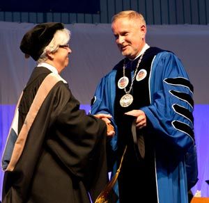 At Saint Joseph's Inauguration, two individuals in academic regalia shake hands—one in a black cap and gown, the other, President James Dlugos, in a blue gown adorned with medals. Saint Joseph's College of Maine