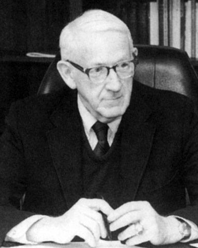 An older gentleman in a suit and tie sits at a desk with a pen in hand, looking thoughtfully to the side, embodying the wisdom that forms the Hart of Campus. Saint Joseph's College of Maine