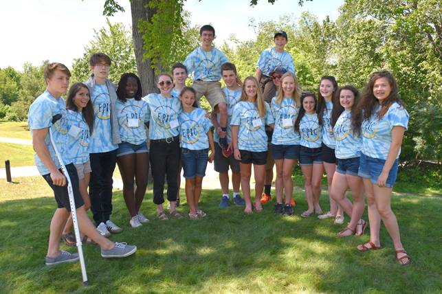 A group of 17 teenagers wearing blue tie-dye shirts stands together outdoors on a sunny day during an E.P.I.C. Retreat at Saint Joseph's College, with one boy holding a crutch and another boy perched on someone’s shoulders. Saint Joseph's College of Maine