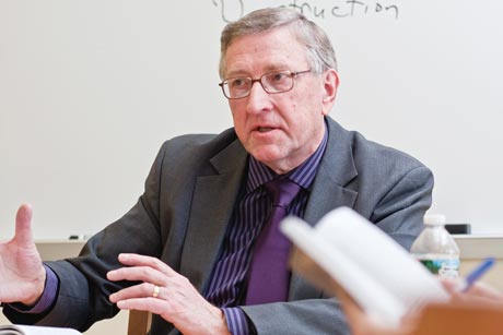 An older man in a suit and glasses, likely a professor, is speaking and gesturing with his hands while sitting at a table. A whiteboard is visible in the background, giving the scene a scholarly atmosphere perfect for this distinguished portrait. Saint Joseph's College of Maine
