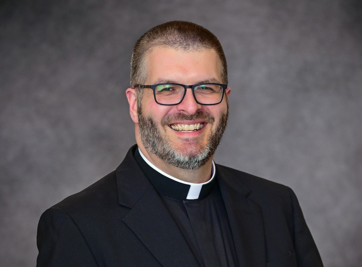 Reverend Patrick Finn, a chaplain at Saint Joseph’s College of Maine, sports short hair, glasses, and a beard. He wears a black clerical collar and suit while smiling against a grey background. Saint Joseph's College of Maine