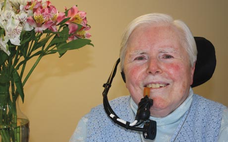 Elderly Sister Anne Fitzpatrick with short white hair uses an assistive communication device. A vase of flowers is in the background. Saint Joseph's College of Maine