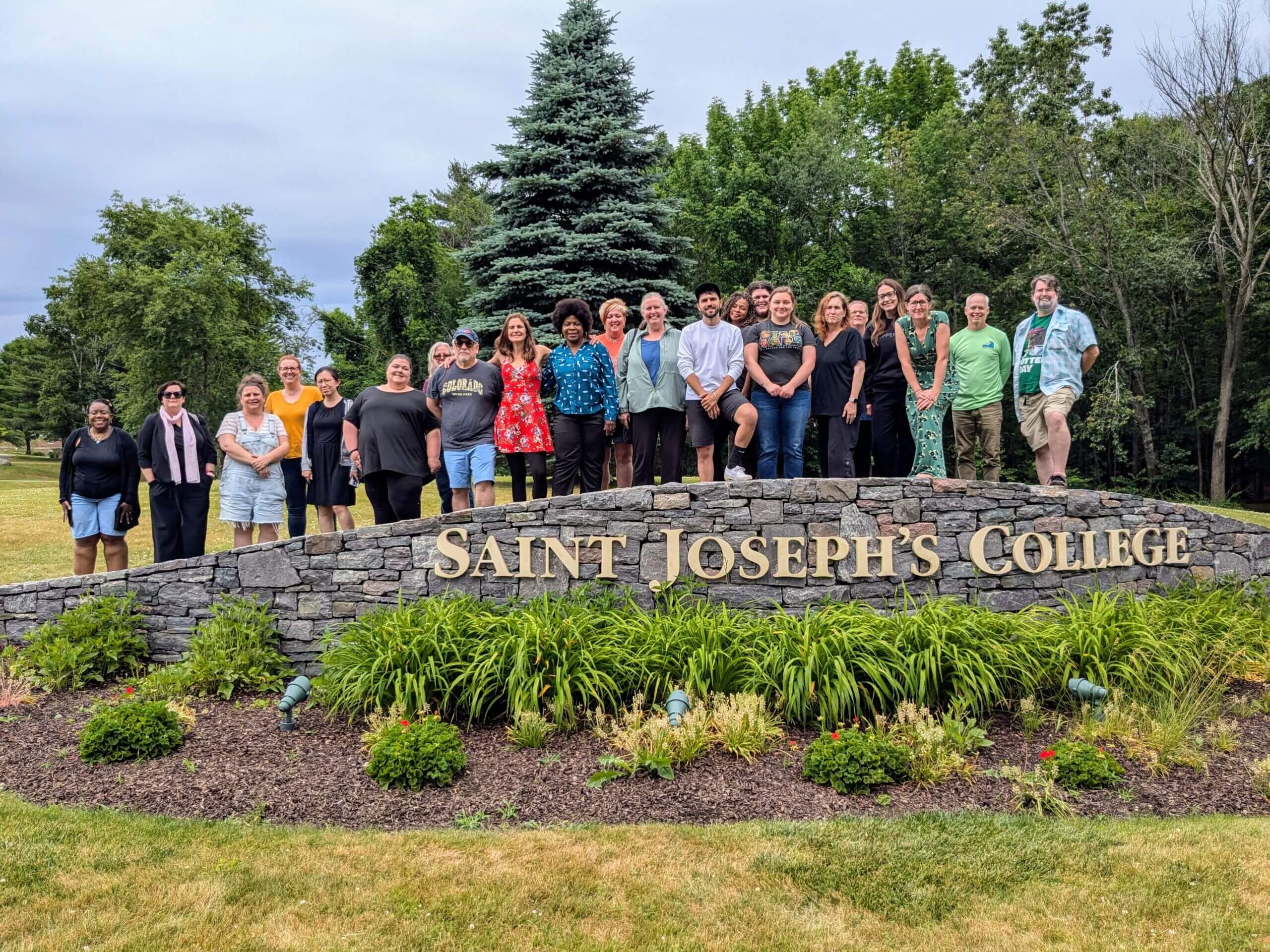 A group of individuals stand together behind a stone sign that reads "Saint Joseph’s College of Maine" in a grassy, tree-lined area, possibly discussing New England Gothic Literature. Saint Joseph's College of Maine