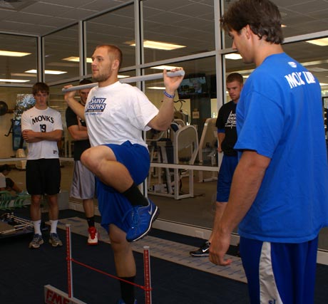 A group of men engage in a gym workout within the Human Performance Laboratory. One man is balancing on one leg while holding a barbell over his shoulders. Others watch or stand near exercise equipment, embodying the principles of the Exercise Science Program. Saint Joseph's College of Maine