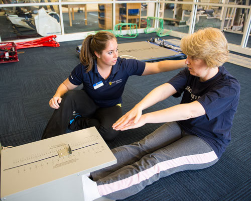 A woman assists another woman in a seated stretching exercise in a gym, using a measurement device on the floor to track health progress. Saint Joseph's College of Maine