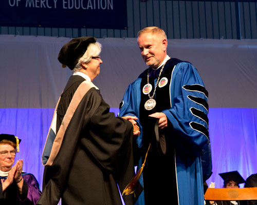 Two individuals in academic regalia shake hands on stage during a formal college event. An audience member in similar attire applauds in the background, marking the inauguration of the 14th president. Saint Joseph's College of Maine