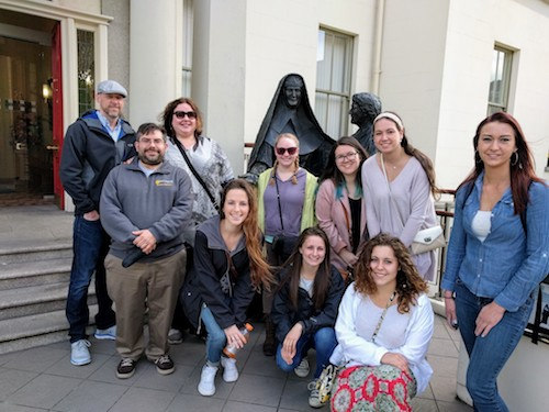 A group of nine people posing in front of a building with two statues behind them during their trip to Ireland. Four are seated on the ground, and five are standing, embarking on a journey of spiritual discovery. Saint Joseph's College of Maine