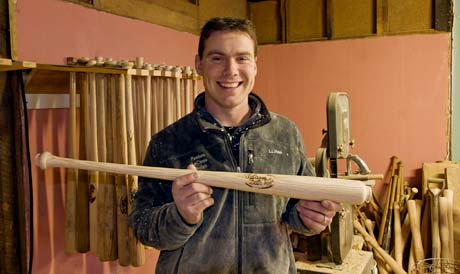 A man smiling and holding a wooden baseball bat in a workshop, with other bats and tools visible in the background. Jesse LaCasse, ’03, proudly showcases his craftsmanship. Saint Joseph's College of Maine