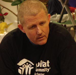 Man with short hair wearing a black Habitat for Humanity T-shirt seated and engaging in conversation, embodying the "theology of the hammer" through his dedication and love for the cause. Saint Joseph's College of Maine