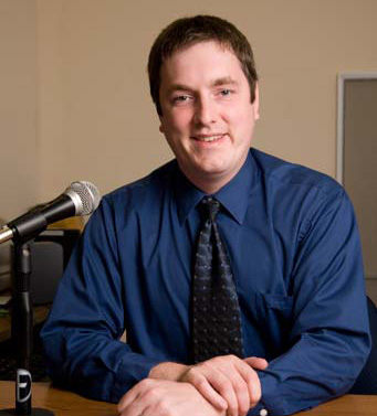 A man in a blue shirt and dark tie sits at a desk with a microphone in front of him, smiling at the camera in a neutral-toned office. This is Justin Sparks, class of 2003. Saint Joseph's College of Maine