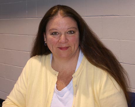 A woman with long brown hair, wearing a yellow cardigan and a white shirt, is sitting against a light-colored brick wall, looking into the camera. Her thoughtful expression suggests she's contemplating a new major in special education. Saint Joseph's College of Maine