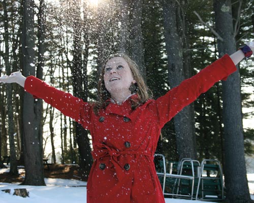 Kathryn Bois, in a red coat, stands in a snowy forest with arms outstretched, looking up as snow falls around her. Saint Joseph's College of Maine