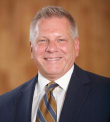 A man with short gray hair is smiling, wearing a dark suit, light shirt, and striped tie. The background is a blurred warm-toned setting. Kenneth Lemanski, Interim President, exudes confidence and professionalism. Saint Joseph's College of Maine
