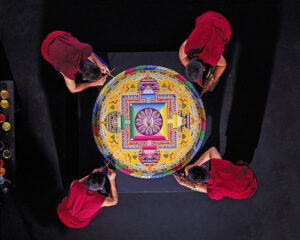 Four Tibetan Buddhist monks in red robes meticulously craft an intricate sand mandala on a black surface at Saint Joseph’s College, viewed from above. Saint Joseph's College of Maine