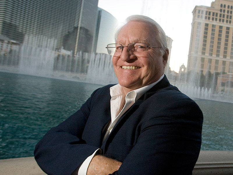 Elderly man with glasses and white hair smiling, standing with arms crossed in front of a body of water and modern high-rise buildings. This portrait captures Larry Messner's confident demeanor, showcasing him as a true leader amidst the urban landscape. Saint Joseph's College of Maine