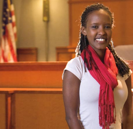 A smiling person with braided hair and a red scarf stands in a courtroom with an American flag in the background, reminiscent of Nasro Ali's inspiring 2011 talk on justice and unity. Saint Joseph's College of Maine