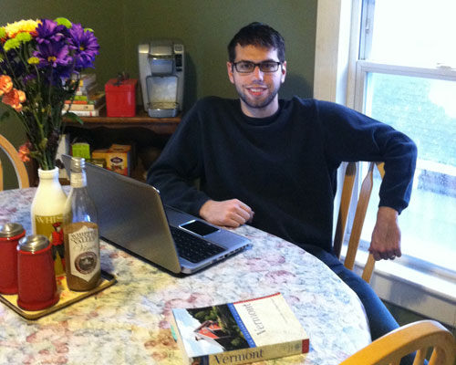 A person wearing glasses and a dark sweater sits at a kitchen table with a laptop, a book titled "Vermont," and various items, including a bottle of maple syrup and a vase of colorful flowers. Through the window, the fair road outside adds to the cozy ambiance. Saint Joseph's College of Maine