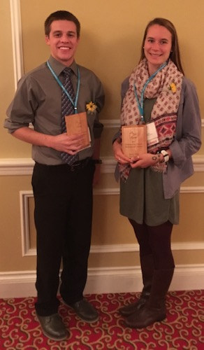 Two people stand side by side, smiling and each holding award plaques for Outstanding Future Professionals. They wear business-casual attire with conference lanyards around their necks. The background features a beige wall and red carpeting, capturing the moment Emily Field is recognized for her achievements in Physical Education. Saint Joseph's College of Maine