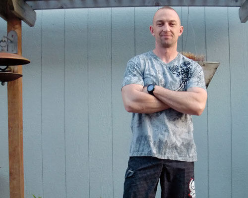 A man with short hair stands with his arms crossed, exuding confidence as the Leader of the Pack. He's wearing a grey graphic T-shirt and black shorts, outdoors near a wooden structure and a grey wall. Saint Joseph's College of Maine