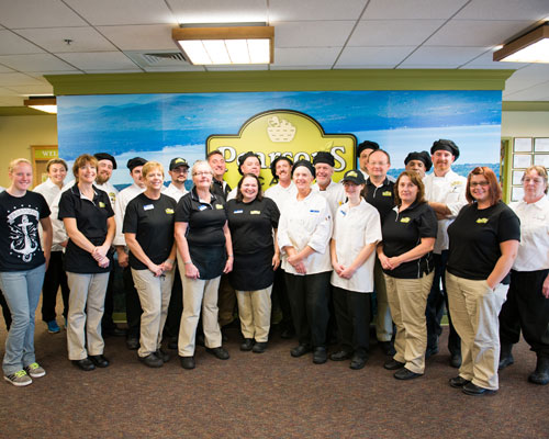 A group of people in uniform stand together posing for a photo inside a building with a sign that reads "Paragon's" in the background, showcasing their local food service unity. Saint Joseph's College of Maine