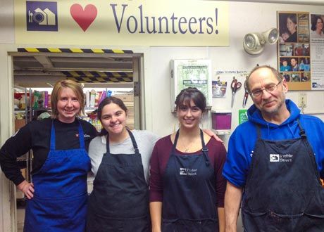 Four people wearing aprons, including faculty volunteers and students, are standing in front of a "♥ Volunteers!" sign inside a room with various supplies and photographs on the walls. Saint Joseph's College of Maine