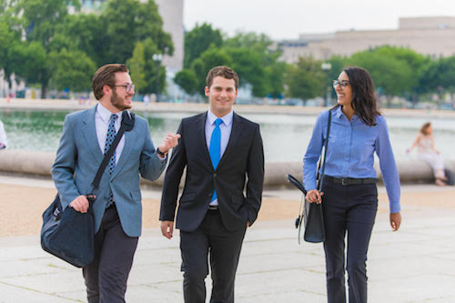 Three people in professional attire walk and talk outdoors near a body of water in an urban park setting, giving the scene a vibe reminiscent of Summer on Capitol Hill. Saint Joseph's College of Maine