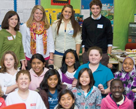 A group of young adults stands behind a group of children in a classroom, participating in a service learning program. The setting appears to be educational, with posters and educational materials visible in the background. Saint Joseph's College of Maine